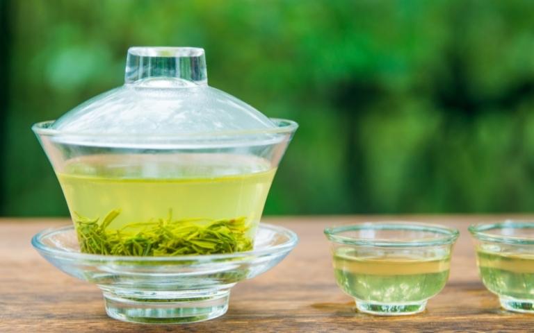 Gaiwan with green tea and tea leaves near two shots of green tea.
