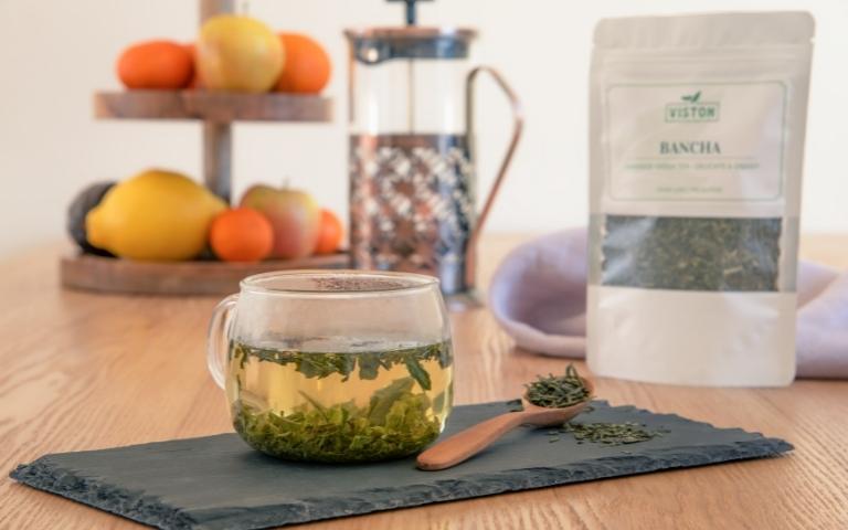An infusion of Viston Japanese bancha tea, wooden tea spoon containing loose-leaf bancha, and a bag of loose-leaf tea in the background.